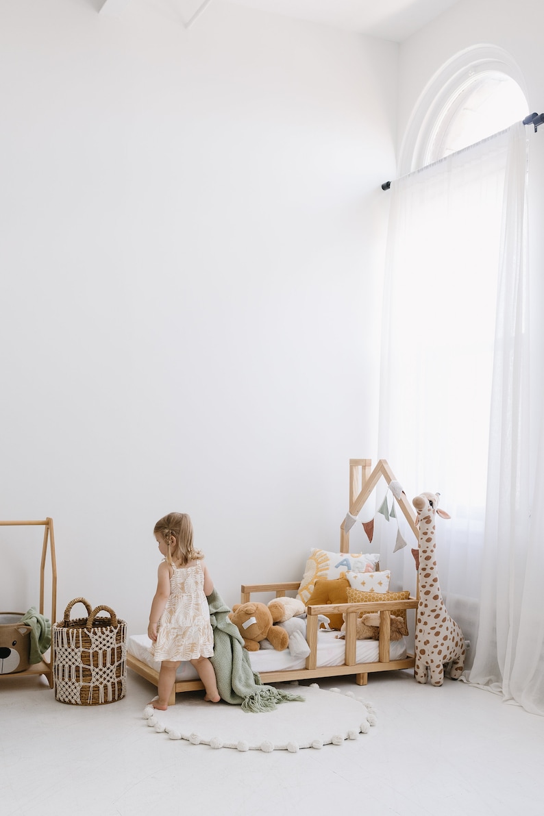 K&ouml;nnte beinhalten: Ein kleines Kind steht in der N&auml;he eines Kinderbettes aus Holz mit einer wei&szlig;en und gr&uuml;nen Decke, einer Stoffgiraffe und verschiedenen bunten Kissen. Das Bett ist mit einem h&ouml;lzernen, hausf&ouml;rmigen Kopfteil und einem wei&szlig;en Pompon-Teppich verziert.