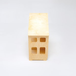 May include: A light-colored wooden bench with a smooth, natural grain finish. The structure features a rectangular top surface and a vertical base with four distinct, square-shaped cutouts arranged in a two-by-two grid pattern on the front side.