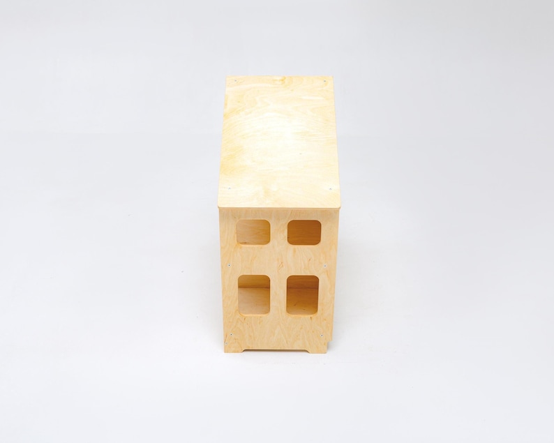 May include: A light-colored wooden bench with a smooth, natural grain finish. The structure features a rectangular top surface and a vertical base with four distinct, square-shaped cutouts arranged in a two-by-two grid pattern on the front side.