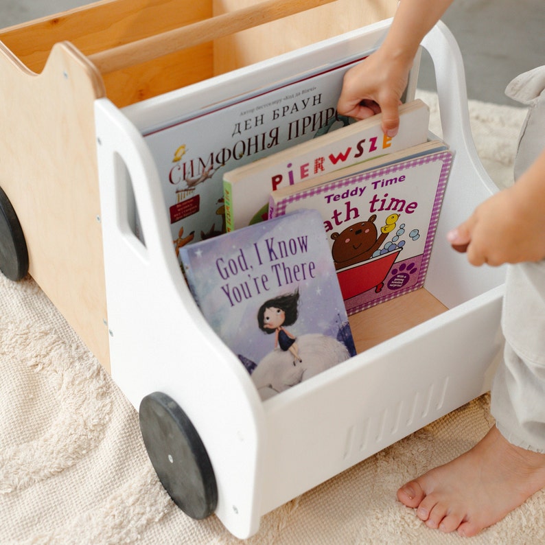 Puede incluir: Un cami&oacute;n de juguete de madera blanco con ruedas negras, lleno de libros infantiles coloridos. Los libros incluyen t&iacute;tulos como "Teddy Time", "God, I Know You're There", y "Pierwsze".