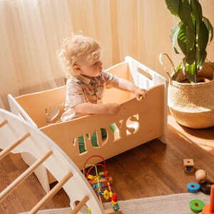 May include: A wooden toy box with the word "TOYS" cut out, a white climbing arch, and a variety of wooden toys. A child is sitting in the toy box. A large woven basket with a plant is in the background.