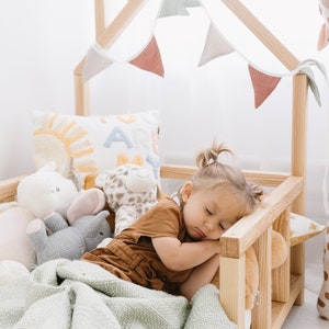 K&ouml;nnte beinhalten: Ein kleines Kind schl&auml;ft in einem h&ouml;lzernen Spielhausbett mit einer gr&uuml;nen Decke, umgeben von Stofftieren. Das Bett hat einen Holzrahmen und einen wei&szlig;en Stoffbaldachin mit einer Schnur aus Stofffahnen.