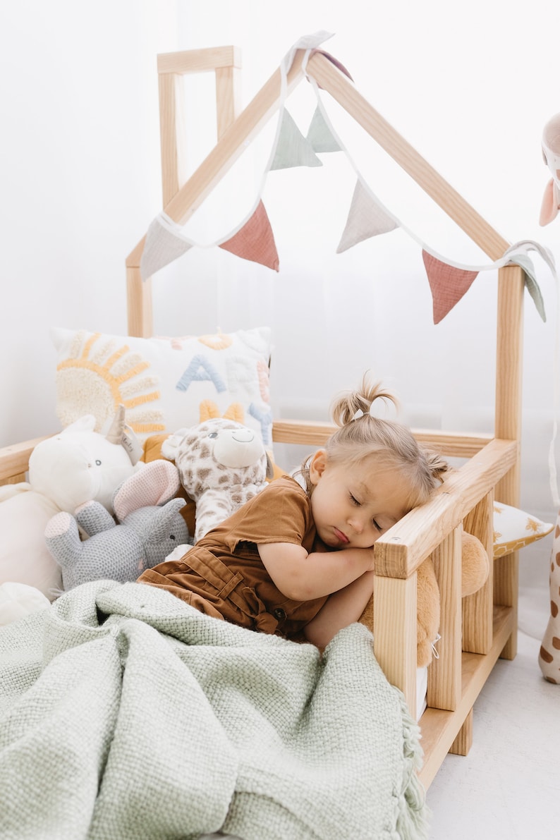 K&ouml;nnte beinhalten: Ein kleines Kind schl&auml;ft in einem h&ouml;lzernen Spielhausbett mit einer gr&uuml;nen Decke, umgeben von Stofftieren. Das Bett hat einen Holzrahmen und einen wei&szlig;en Stoffbaldachin mit einer Schnur aus Stofffahnen.