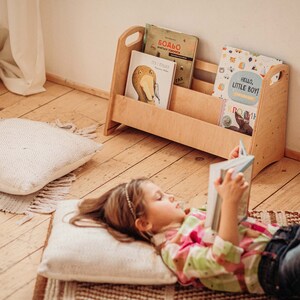 Puede incluir: Una ni&ntilde;a peque&ntilde;a est&aacute; tumbada en una alfombra leyendo un libro. Est&aacute; rodeada de almohadas y una estanter&iacute;a de madera llena de libros infantiles. La estanter&iacute;a tiene dos estantes y un asa en la parte superior. Los libros de la estanter&iacute;a tienen cubiertas coloridas con ilustraciones de animales y texto en diferentes idiomas. La alfombra es marr&oacute;n y blanca con un patr&oacute;n geom&eacute;trico. Las almohadas son blancas y esponjosas.