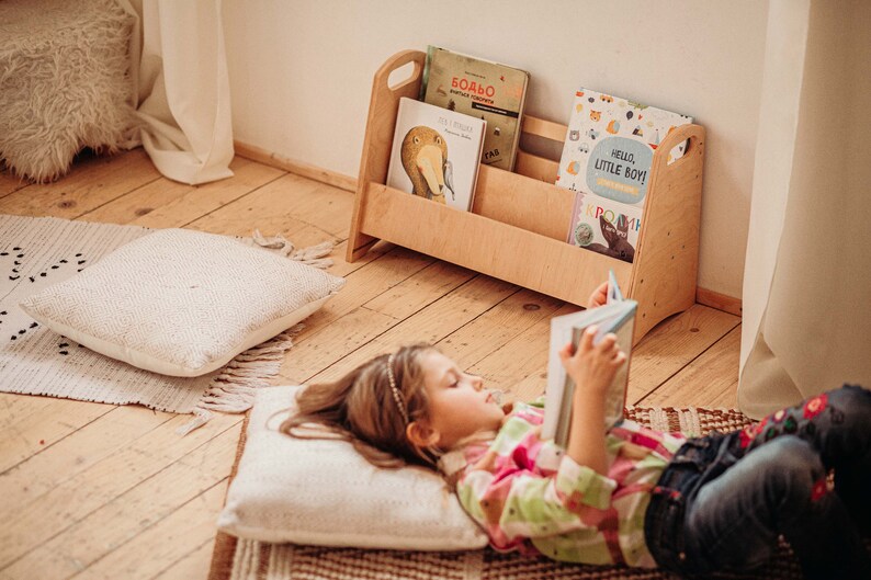 Puede incluir: Una ni&ntilde;a peque&ntilde;a est&aacute; tumbada en una alfombra leyendo un libro. Est&aacute; rodeada de almohadas y una estanter&iacute;a de madera llena de libros infantiles. La estanter&iacute;a tiene dos estantes y un asa en la parte superior. Los libros de la estanter&iacute;a tienen cubiertas coloridas con ilustraciones de animales y texto en diferentes idiomas. La alfombra es marr&oacute;n y blanca con un patr&oacute;n geom&eacute;trico. Las almohadas son blancas y esponjosas.