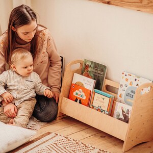 Puede incluir: Una estanter&iacute;a de libros de madera con un asa a cada lado, llena de libros infantiles coloridos. Un ni&ntilde;o peque&ntilde;o est&aacute; sentado en el suelo con un adulto, mirando los libros.