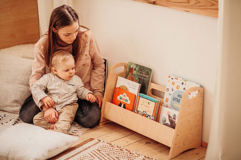 Puede incluir: Una estanter&iacute;a de libros de madera con un asa a cada lado, llena de libros infantiles coloridos. Un ni&ntilde;o peque&ntilde;o est&aacute; sentado en el suelo con un adulto, mirando los libros.