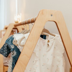 May include: A light-colored wooden clothing rack with an A-frame design, featuring a horizontal rod holding several hangers with various garments, including a white top with a small cherry print and ruffled sleeves, and a blue patterned dress.