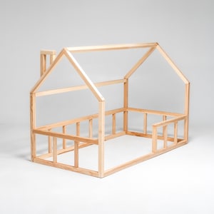 May include: A wooden house-shaped bed frame for a child. The frame is made of natural wood and has a simple design. It features a raised headboard and footboard, as well as a small chimney on the roof.