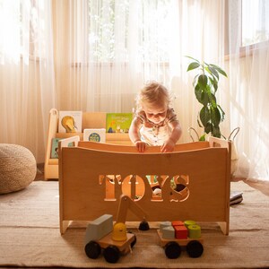 May include: A light-coloured wooden toy box with the word "TOYS" cut out on the front. A child is playing with the toys inside. A wooden toy excavator and a toy truck are in front of the box. Bookshelves and a woven pouf are in the background.