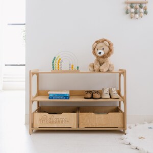 May include: A wooden shelf unit with two shelves and two drawers. The top shelf has a rainbow-colored abacus and a stuffed lion. The bottom shelf has two books and a pair of shoes. The drawers are labeled "Adam".