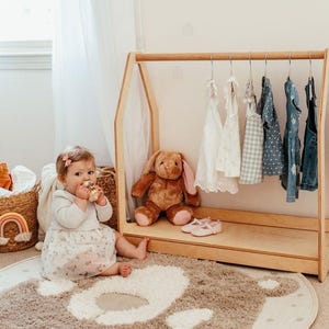 May include: A light-colored wooden A-frame clothing rack featuring a horizontal rod with six hangers holding various children's garments, including white dresses, a checkered top, and blue denim overalls, with a brown plush rabbit toy inside.