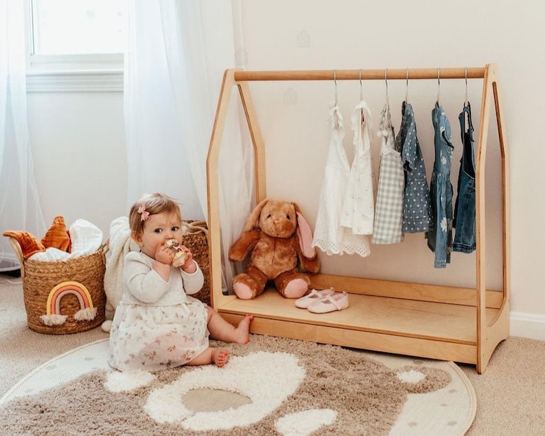 May include: A light-colored wooden A-frame clothing rack featuring a horizontal rod with six hangers holding various children's garments, including white dresses, a checkered top, and blue denim overalls, with a brown plush rabbit toy inside.