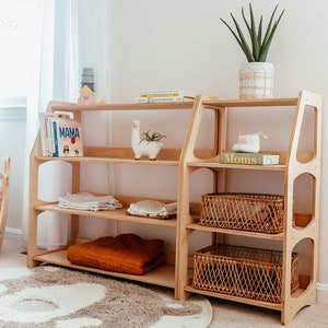 May include: A light-colored wooden shelving unit with multiple tiers, featuring a wide section with three open shelves and a narrower section with three shelves, two of which contain woven baskets. The unit has rounded side cutouts.