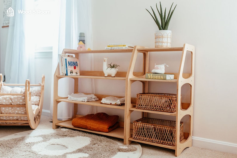 May include: A light-colored wooden shelving unit with multiple tiers, featuring a wide section with three open shelves and a narrower section with three shelves, two of which contain woven baskets. The unit has rounded side cutouts.