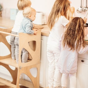 May include: Three children stand at a kitchen counter, two using a wooden learning tower and step stool to reach the counter. The tower is made of light wood with a simple design. The step stool is also made of light wood and has a simple design.
