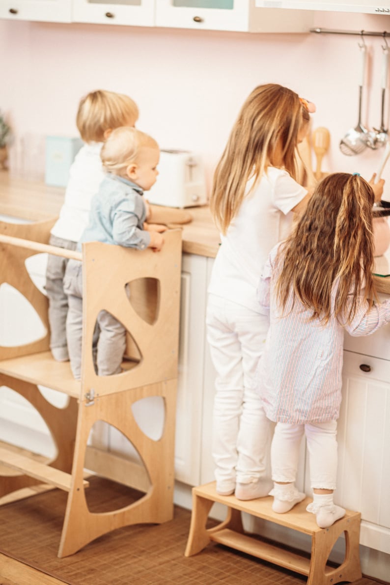 May include: Three children stand at a kitchen counter, two using a wooden learning tower and step stool to reach the counter. The tower is made of light wood with a simple design. The step stool is also made of light wood and has a simple design.