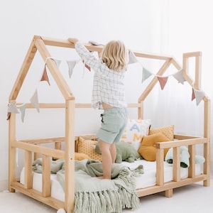 May include: A natural wood-framed house bed with a white mattress, decorated with colorful pillows, a green blanket, and a banner of triangular flags. A child is standing on the bed, reaching for the banner. The bed is a cozy and inviting space for a child.
