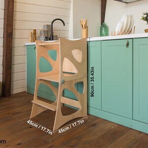 May include: A wooden step stool with a unique design, featuring rounded edges and a cutout pattern. It is 17.7 inches tall and 17.7 inches wide. The stool is shown in a kitchen setting, next to a light green cabinet.