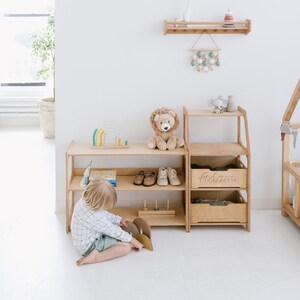 May include: A wooden shelf unit with two shelves and a drawer. The drawer is labeled "Adam". There are shoes, books, and toys on the shelves. A child is playing with a wooden toy on the floor in front of the shelf.