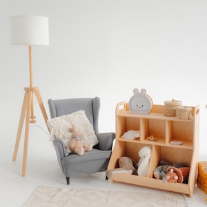 K&ouml;nnte beinhalten: Ein helles Holzspielzeugregal mit sechs F&auml;chern, ein grauer Sessel mit einem wei&szlig;en H&auml;kelkissen, eine Stehlampe mit einem wei&szlig;en Lampenschirm und Stofftiere. Das Regal hat ein wei&szlig;es Kaninchen-Gesicht auf der Vorderseite.
