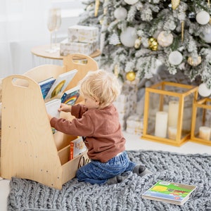 K&ouml;nnte beinhalten: Ein Kind greift nach einem Buch in einem h&ouml;lzernen B&uuml;cherregal mit mehreren Regalbrettern. Das B&uuml;cherregal steht in einem Raum mit einem wei&szlig;en Teppich und einem Weihnachtsbaum, der mit wei&szlig;en und goldenen Kugeln geschm&uuml;ckt ist.