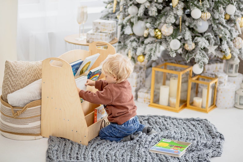 K&ouml;nnte beinhalten: Ein Kind greift nach einem Buch in einem h&ouml;lzernen B&uuml;cherregal mit mehreren Regalbrettern. Das B&uuml;cherregal steht in einem Raum mit einem wei&szlig;en Teppich und einem Weihnachtsbaum, der mit wei&szlig;en und goldenen Kugeln geschm&uuml;ckt ist.
