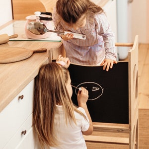 May include: Two children, one drawing on a chalkboard easel and the other holding a pink kettle and a spoon. The easel is made of wood and has a black chalkboard surface. The children are in a kitchen setting.