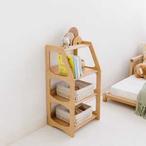 Op de afbeelding: Een houten boekenkast met vier planken, met drie geweven manden en een verzameling boeken. De boekenkast is in een lichte houttint en heeft een uniek gebogen ontwerp.