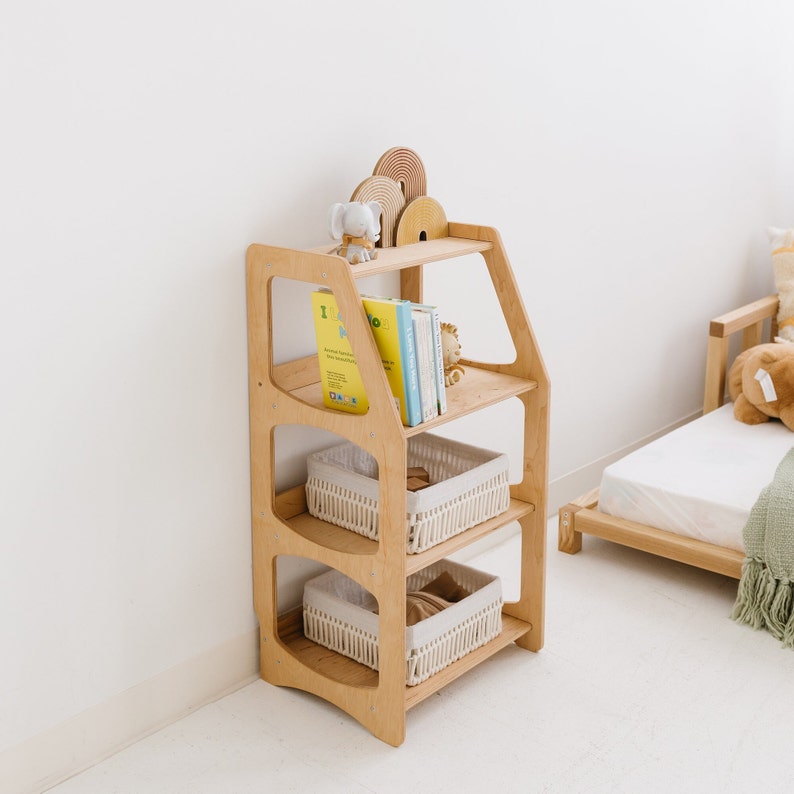 Op de afbeelding: Een houten boekenkast met vier planken, met drie geweven manden en een verzameling boeken. De boekenkast is in een lichte houttint en heeft een uniek gebogen ontwerp.