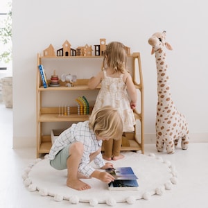 Pode incluir: Duas crian&ccedil;as brincam em um quarto com uma estante de madeira, uma girafa de pel&uacute;cia e um tapete branco e felpudo. A estante tem brinquedos e livros nas prateleiras. As crian&ccedil;as est&atilde;o olhando para um livro.