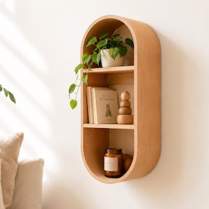 May include: A light-colored wooden wall shelf with an oval shape and three shelves. The top shelf holds a potted plant. The middle shelf has a book and a wooden stacking toy. The bottom shelf has a candle in a glass jar.