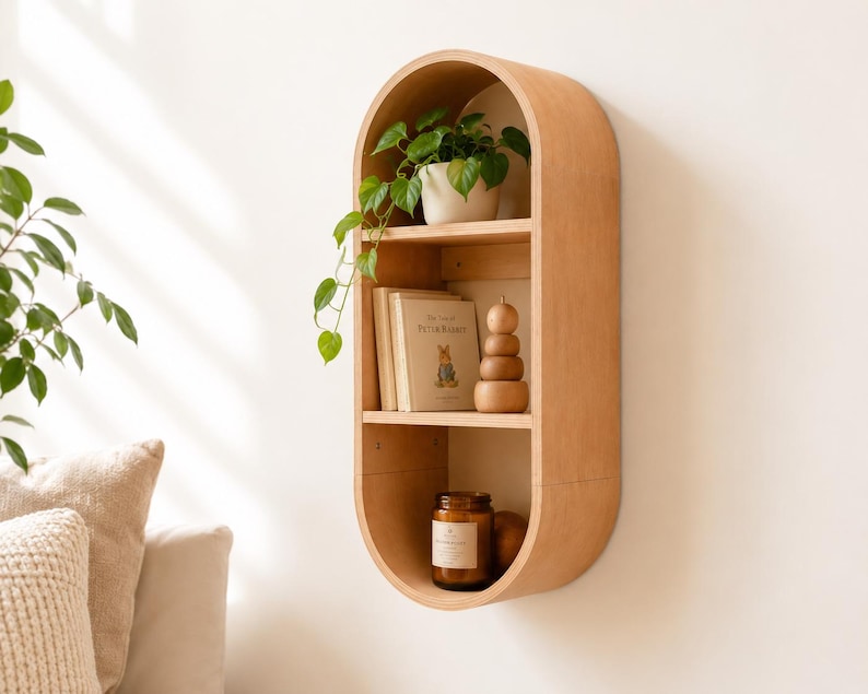 May include: A light-colored wooden wall shelf with an oval shape and three shelves. The top shelf holds a potted plant. The middle shelf has a book and a wooden stacking toy. The bottom shelf has a candle in a glass jar.