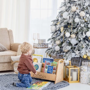 K&ouml;nnte beinhalten: Ein Holz-Buchregal mit dem personalisierten Namen "Adam" auf der Vorderseite. Das Regal ist mit Kinderb&uuml;chern gef&uuml;llt und steht auf einem grauen Teppich. Ein kleines Kind kniet vor dem Regal und betrachtet die B&uuml;cher. Im Hintergrund steht ein wei&szlig;er Weihnachtsbaum mit goldenen und wei&szlig;en Ornamenten.