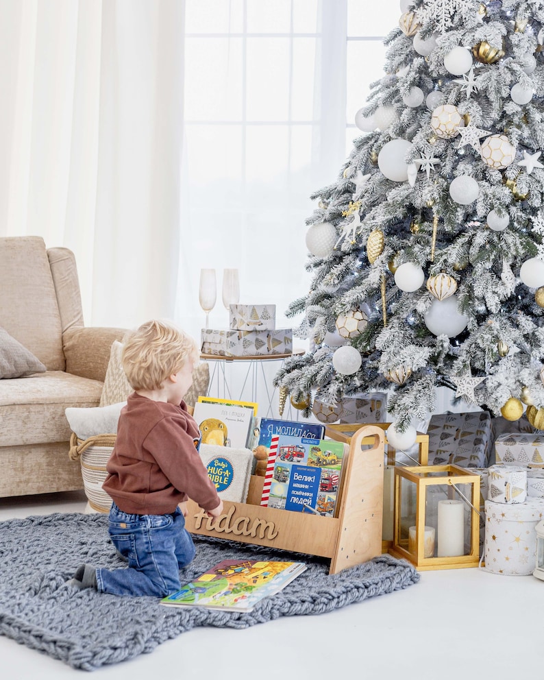 K&ouml;nnte beinhalten: Ein Holz-Buchregal mit dem personalisierten Namen "Adam" auf der Vorderseite. Das Regal ist mit Kinderb&uuml;chern gef&uuml;llt und steht auf einem grauen Teppich. Ein kleines Kind kniet vor dem Regal und betrachtet die B&uuml;cher. Im Hintergrund steht ein wei&szlig;er Weihnachtsbaum mit goldenen und wei&szlig;en Ornamenten.