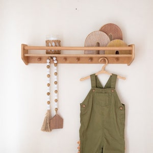 Pu&ograve; includere: Uno scaffale in legno con una finitura in legno chiaro, con un arcobaleno decorativo in legno, un barattolo di vetro e una ghirlanda di perline. Una salopette verde &egrave; appesa a un gancio di legno sullo scaffale.
