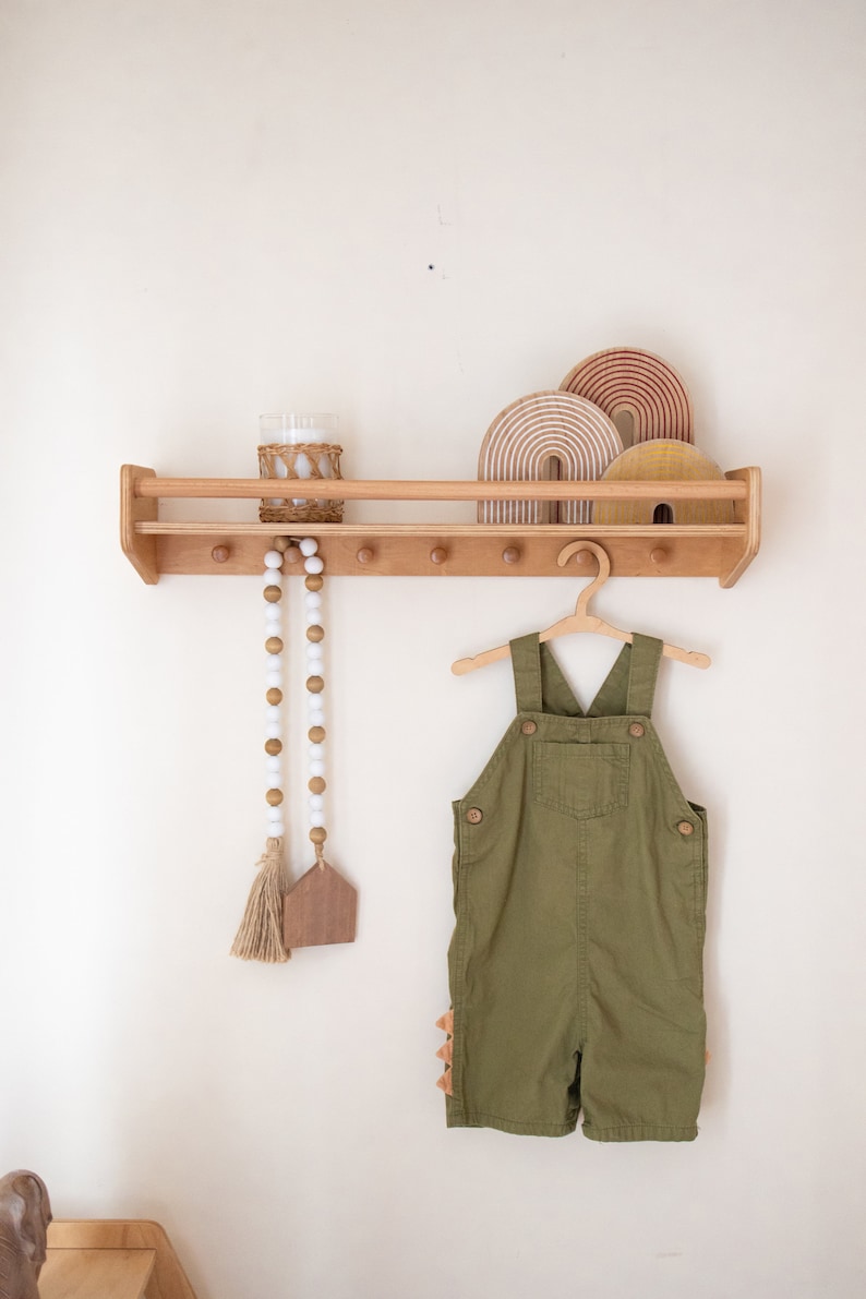 Pu&ograve; includere: Uno scaffale in legno con una finitura in legno chiaro, con un arcobaleno decorativo in legno, un barattolo di vetro e una ghirlanda di perline. Una salopette verde &egrave; appesa a un gancio di legno sullo scaffale.