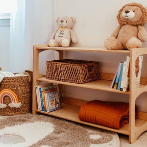 K&ouml;nnte beinhalten: Ein dreist&ouml;ckiges B&uuml;cherregal aus hellem Holz mit einem braunen geflochtenen Korb, mehreren Kinderb&uuml;chern und einer gefalteten rostfarbenen Decke. Obenauf sitzen ein beiger Teddyb&auml;r mit dem Text My first bear und ein brauner Pl&uuml;schl&ouml;we.