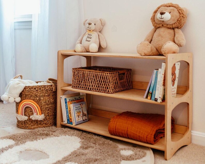 K&ouml;nnte beinhalten: Ein dreist&ouml;ckiges B&uuml;cherregal aus hellem Holz mit einem braunen geflochtenen Korb, mehreren Kinderb&uuml;chern und einer gefalteten rostfarbenen Decke. Obenauf sitzen ein beiger Teddyb&auml;r mit dem Text My first bear und ein brauner Pl&uuml;schl&ouml;we.