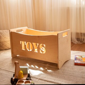 May include: A light-coloured wooden toy box with the word "TOYS" cut out on the front. The box has handles on each side and is sitting on a rug. A wooden toy truck is in front of the box.