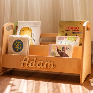 May include: A wooden bookshelf with a nameplate that reads "Adam" and is filled with children's books. The bookshelf has a handle on each side. The books are in Russian and English.