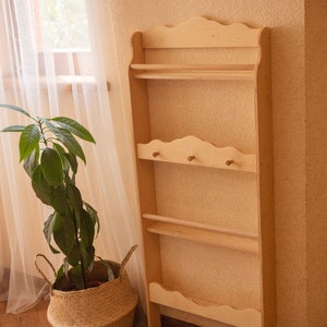 May include: A tall, unfinished wooden shelf unit with multiple shelves and a decorative top. The unit has a natural wood color and is designed for storage or display. A plant in a woven basket sits nearby.