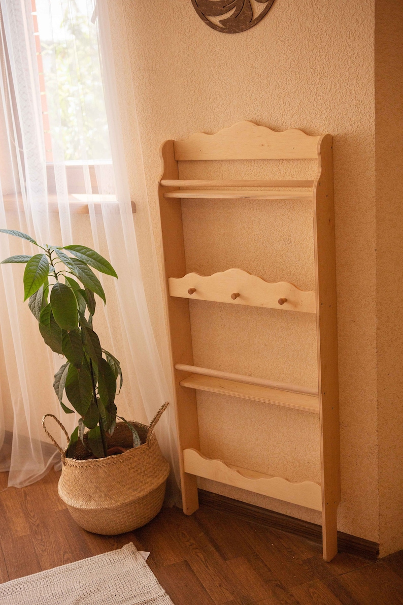 May include: A tall, unfinished wooden shelf unit with multiple shelves and a decorative top. The unit has a natural wood color and is designed for storage or display. A plant in a woven basket sits nearby.