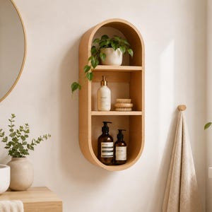 May include: A light wood, oval-shaped wall shelf with three levels, holding a small potted plant, a bottle of lotion, a wooden brush, and two brown bottles. The shelf is mounted on a white wall, with a gold-framed mirror and a sink visible.