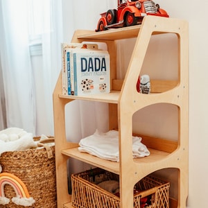 May include: A light-colored wooden bookshelf with three tiers. The top shelf holds a red toy truck. The middle shelf contains a row of children's books, including one titled DADA by Jimmy Fallon, and a folded white garment. A woven basket sits on the bottom shelf.