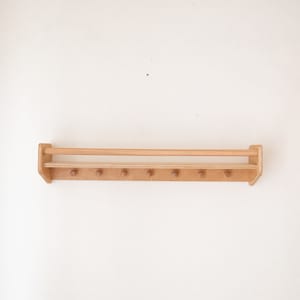 Pu&ograve; includere: Un ripiano a muro in legno con una barra superiore e pi&ugrave; pioli per appendere oggetti. Il ripiano &egrave; in legno marrone chiaro e ha una finitura naturale.