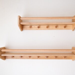 May include: Two wooden wall shelves with a rail and hooks. Each shelf has a natural wood finish and is mounted on a white wall.