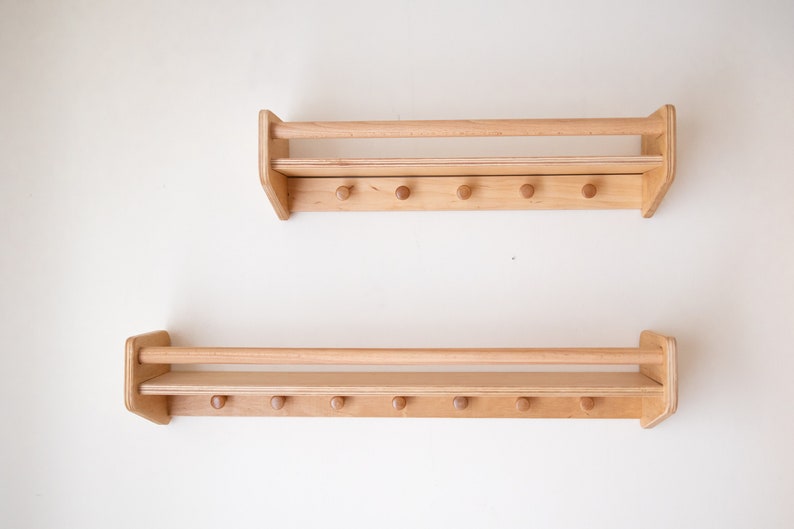 May include: Two wooden wall shelves with a rail and hooks. Each shelf has a natural wood finish and is mounted on a white wall.