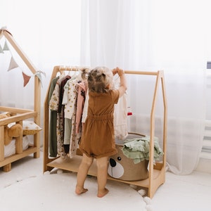 Op de afbeelding: Een houten kledingrek met een bruine en witte beren-vormige opbergmand. Een jong kind staat voor het rek en houdt een bruine jurk vast. Het rek is gevuld met verschillende kledingstukken, waaronder een groene jurk, een roze jurk en een blauwe jurk.