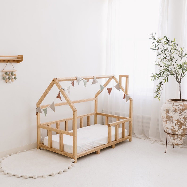 May include: A natural wood-framed house bed with a white mattress. The bed frame has a house-shaped roof and a low profile. A garland of pastel-colored pennants hangs from the roof. A white pom-pom rug sits on the floor.